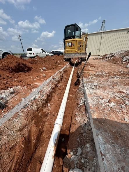 Mini excavator over an open trench with new white sewer pipe laid at a commercial site