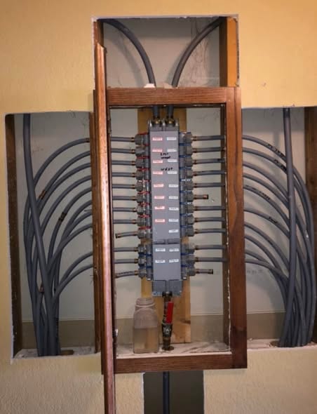 Original gray plastic water manifold exposed in a wall opening, supply lines fanned out through the framing on both sides