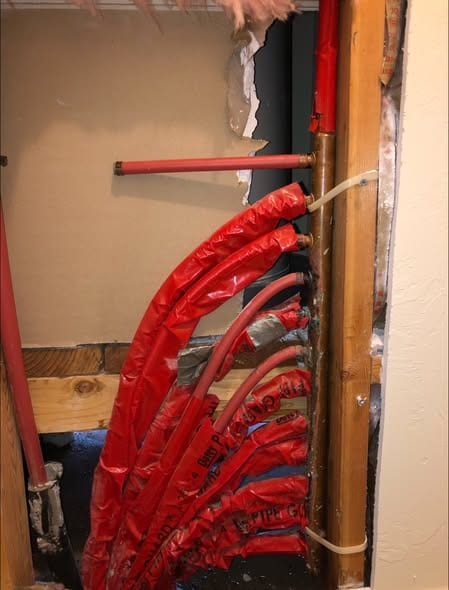 Bundle of insulated red PEX supply lines routed up a wall chase beside a copper stub during a repipe