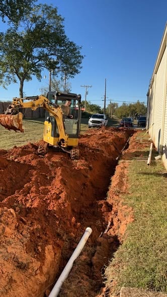 Mini excavator opening a sewer trench along the side of a commercial building