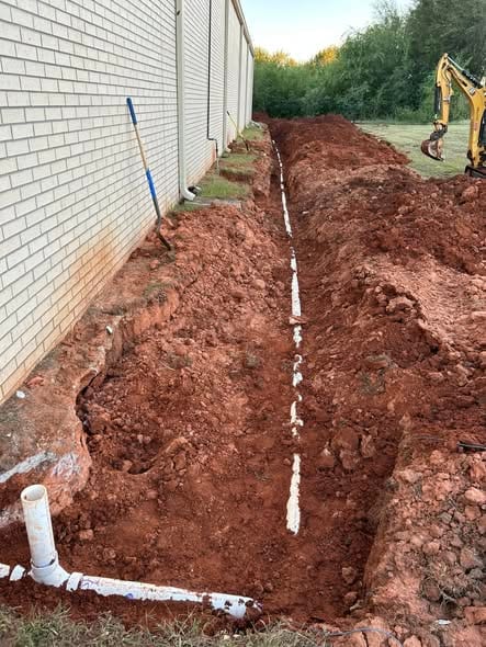 Open trench with a new sewer line running along the side of a commercial building, excavator at the far end
