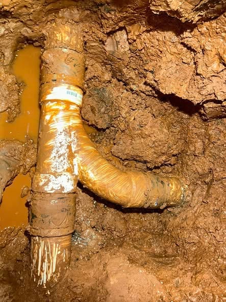 Repaired PVC drain fittings in a wet red-clay excavation beneath the slab